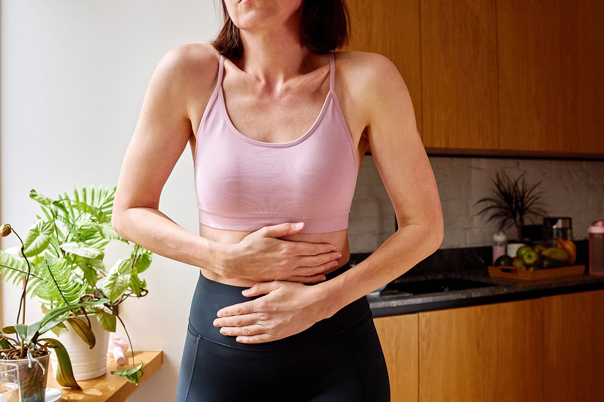 A woman wearing activewear is standing in a kitchen, with her hands on her stomach to indicate pain