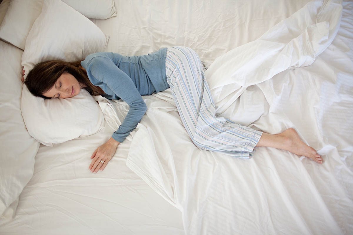 A person is peacefully sleeping on a bed with white bedding, wearing a blue long-sleeve shirt and striped pajama pants.