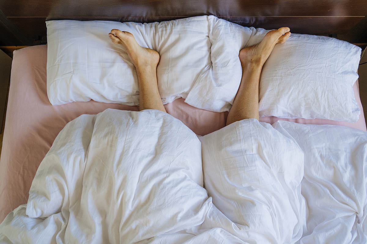A pair of legs are sticking out from under a white duvet, resting on white pillows.
