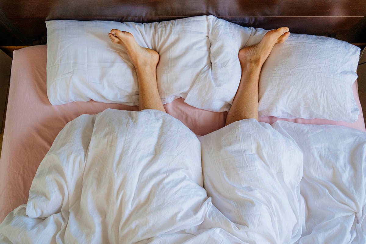 A pair of legs are sticking out from under a white duvet, resting on white pillows.