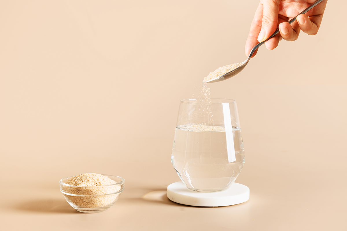 A hand adding a spoonful of psyllium fibre to a glass of water on beige background.