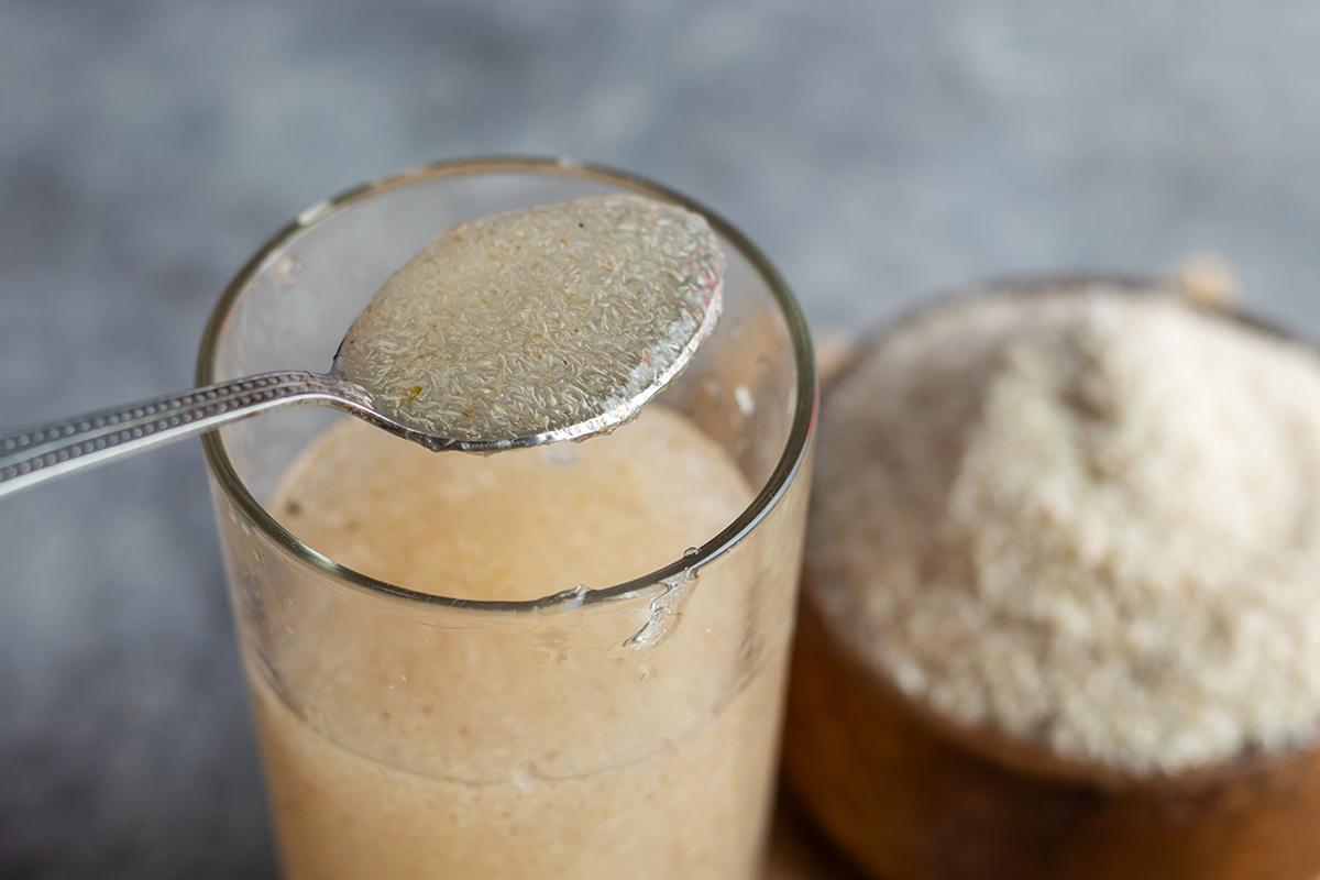 A spoon containing psyllium husk is held above a glass of orange-coloured liquid.