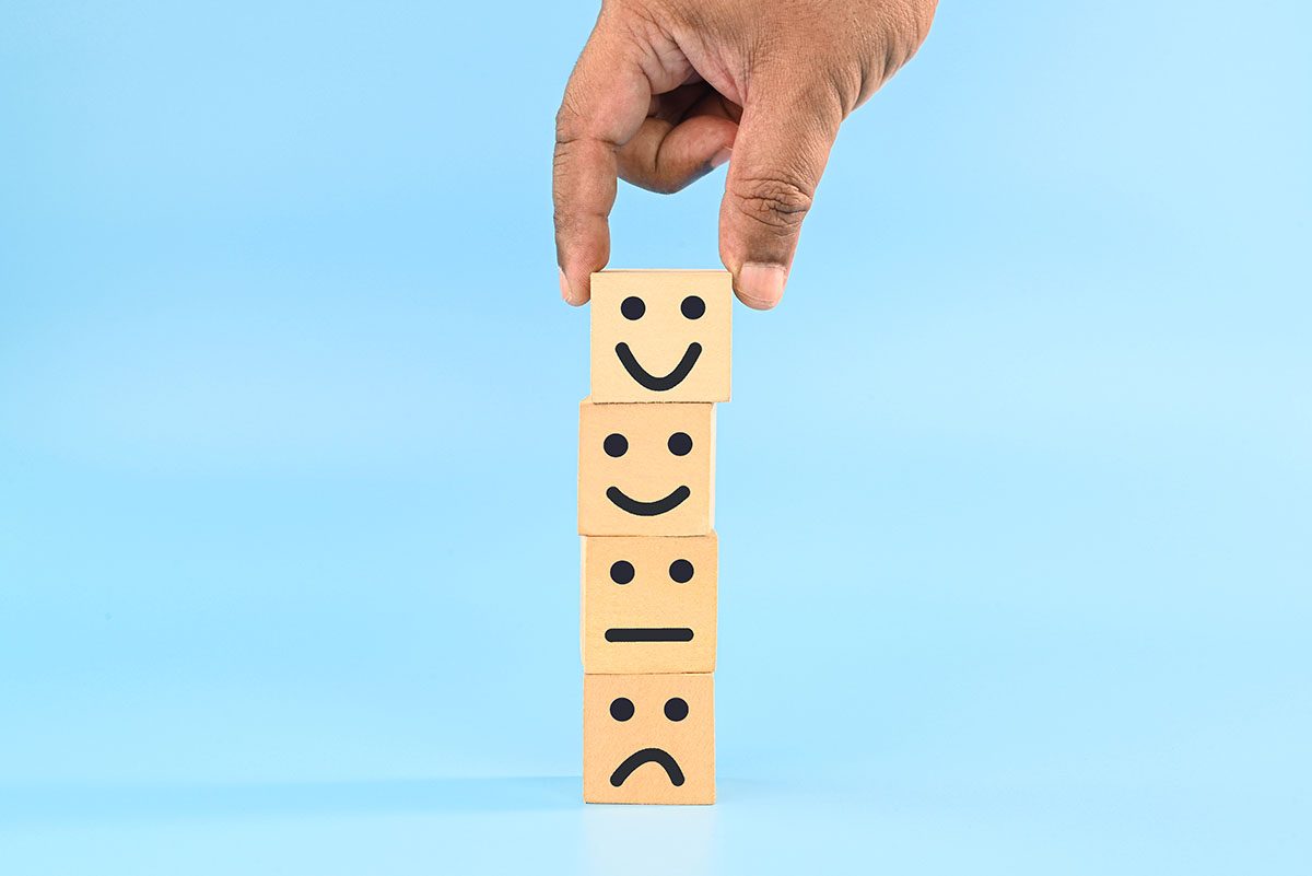 A person stacks wooden tiles, each marked with a happy or sad smiley face, against a blue background