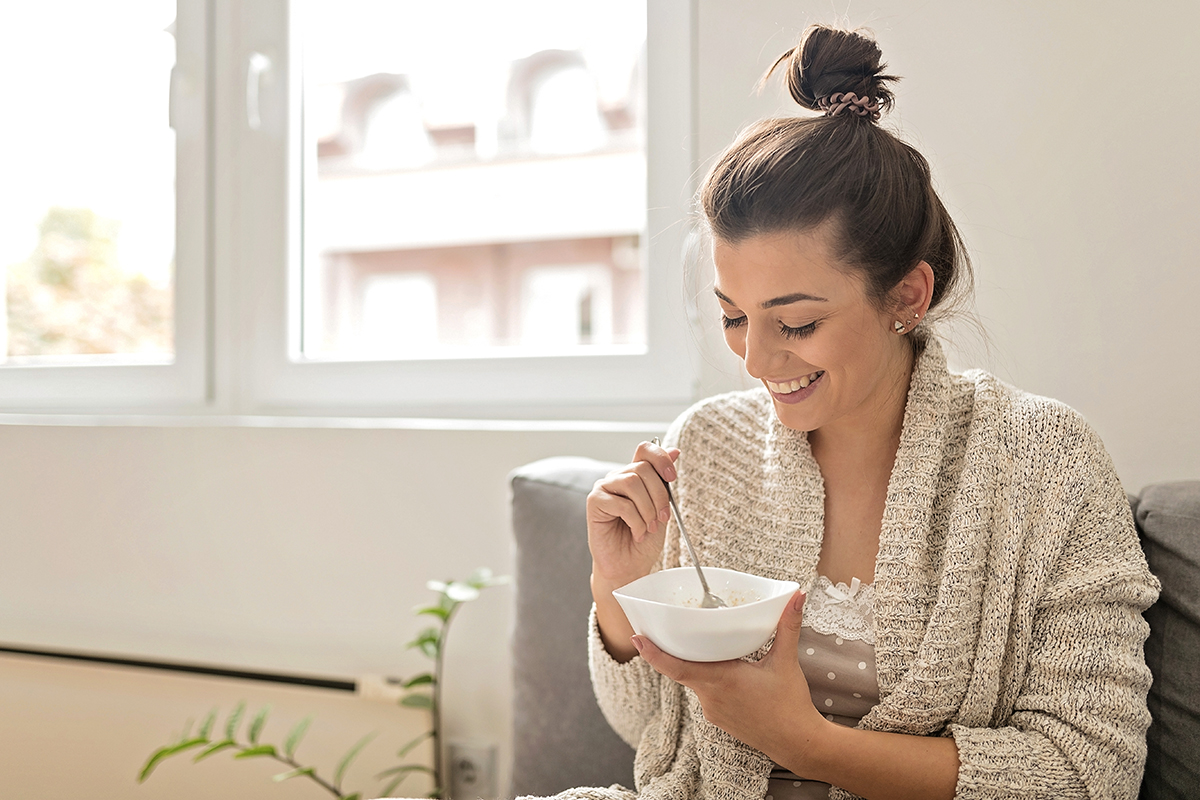 A woman wearing a knitted cardigan is holding a bowl of porridge and smiling.