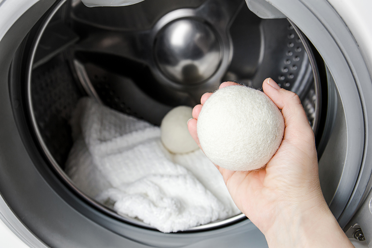 A hand holds a dryer ball in front an open washing machine that has laundry and another dryer ball inside.