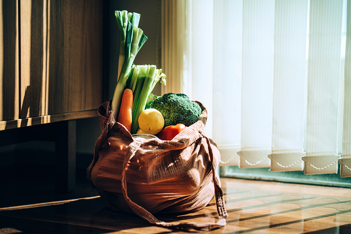 A brown reusable shopping bag filled with vegetables is set on the floor next to white blinds.