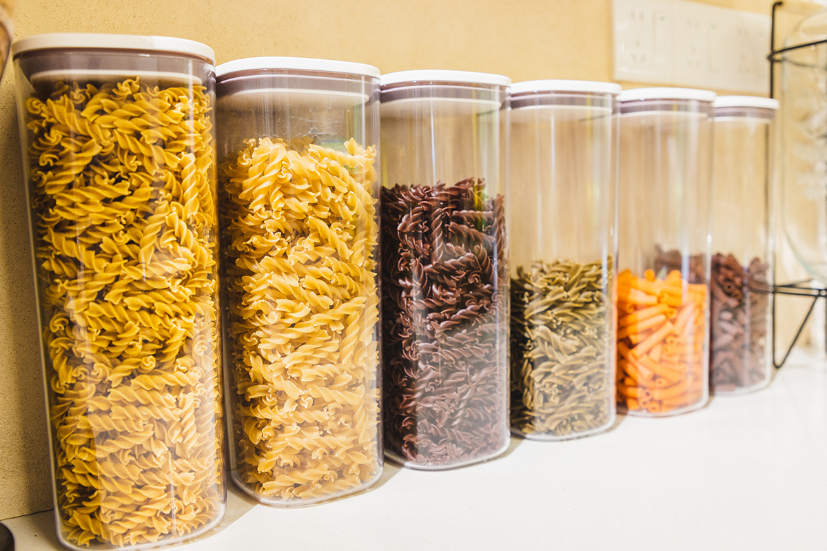 Plastic containers filled with different types of pasta are lined up on a white surface.