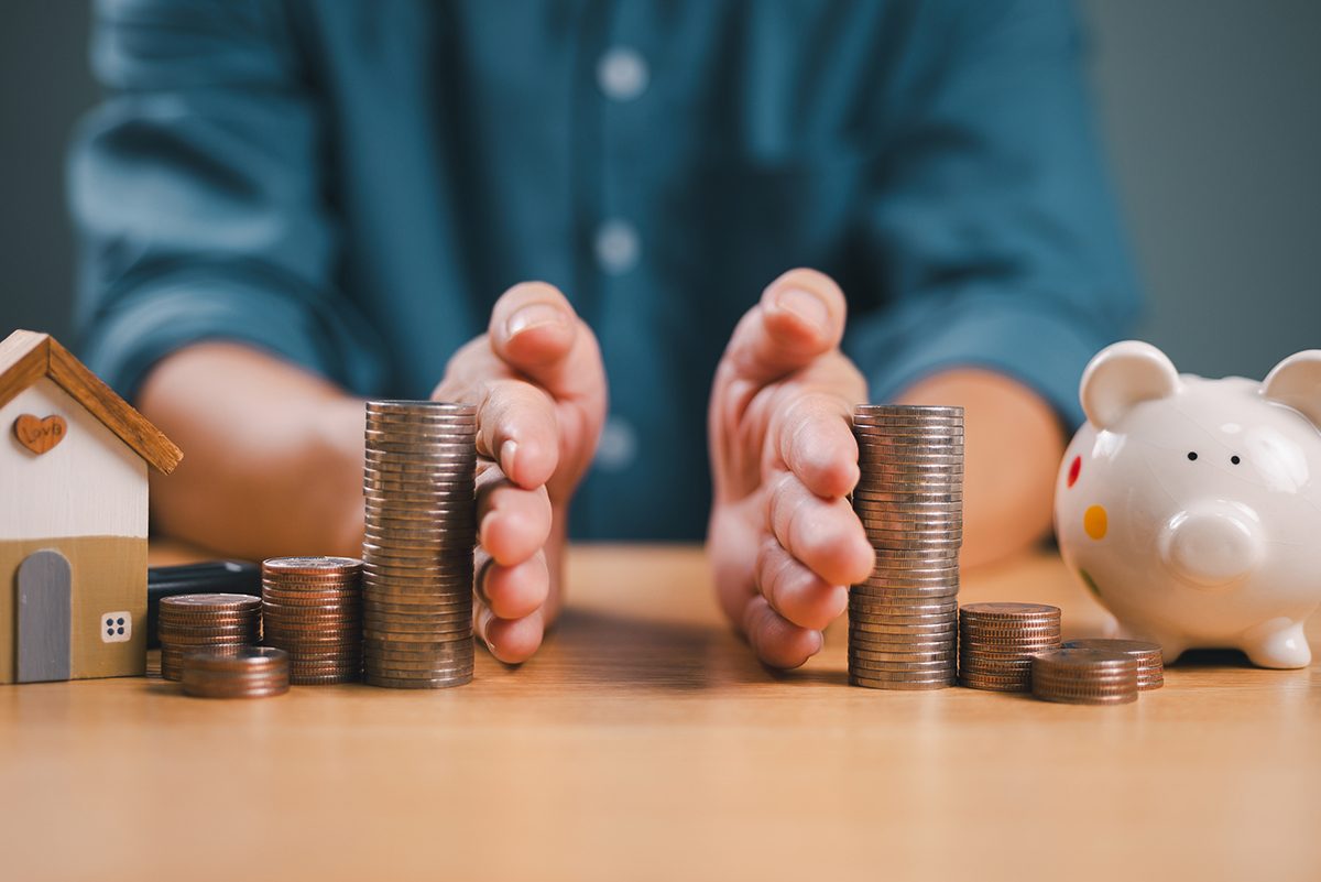 Hands divide stacks of coins, with a miniature house on one side and a piggy bank on the other