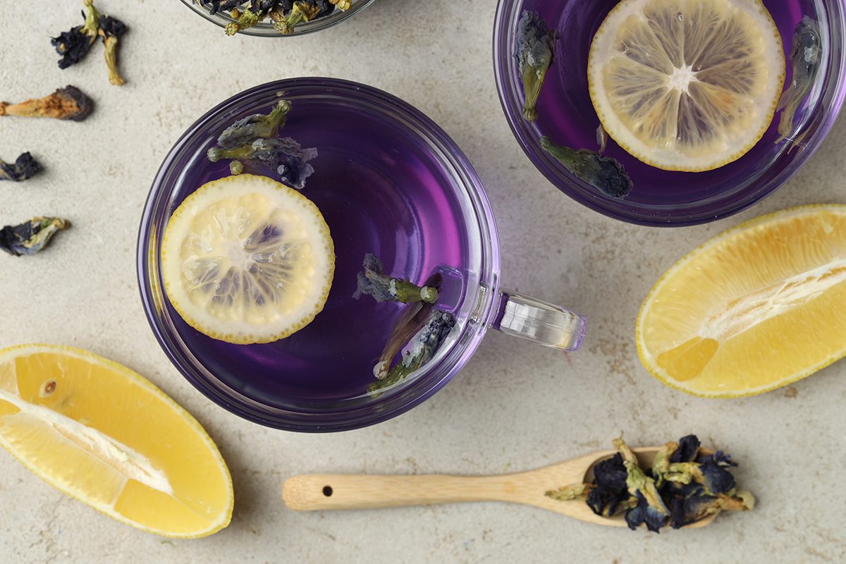 Two glass mugs filled with purple butterfly pea flower tea, garnished with lemon slices and surrounded by fresh lemon wedges and dried butterfly pea flowers