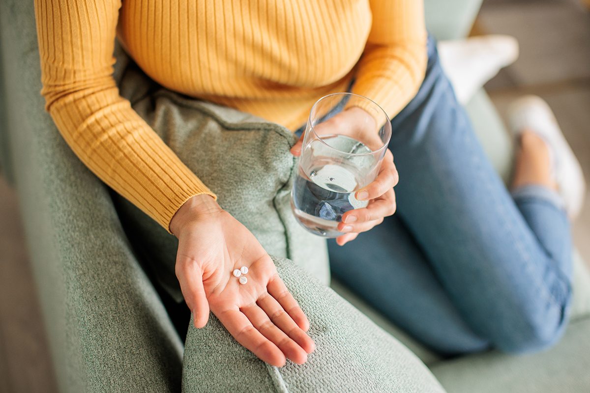 A woman sitting on a sofa holds a glass of water in one hand and has three pills in the other