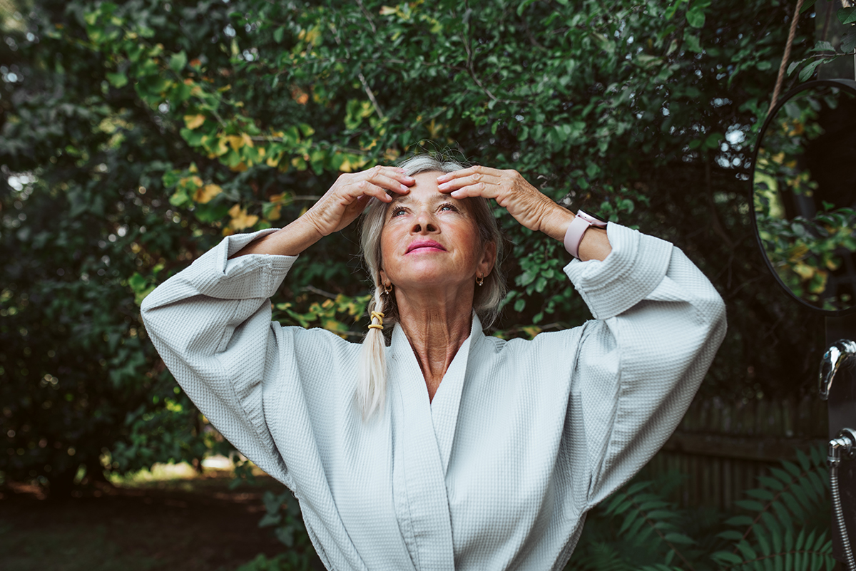 A mature woman is standing outdoors softly touching her forehead