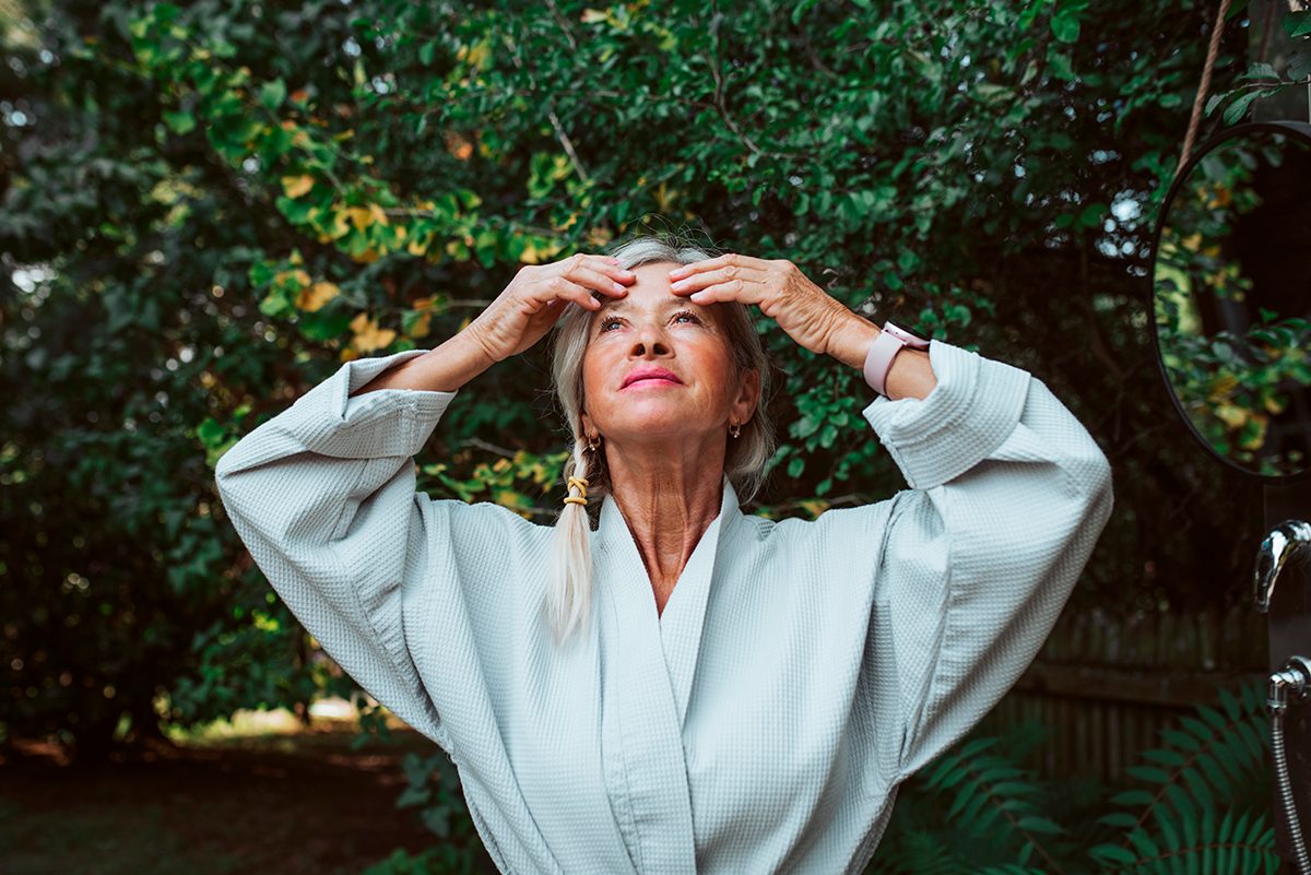 A mature woman is standing outdoors softly touching her forehead