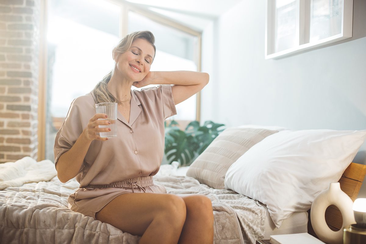 A woman wearing pyjamas is sitting on a bed with her eyes closed, holding a glass of water in one hand