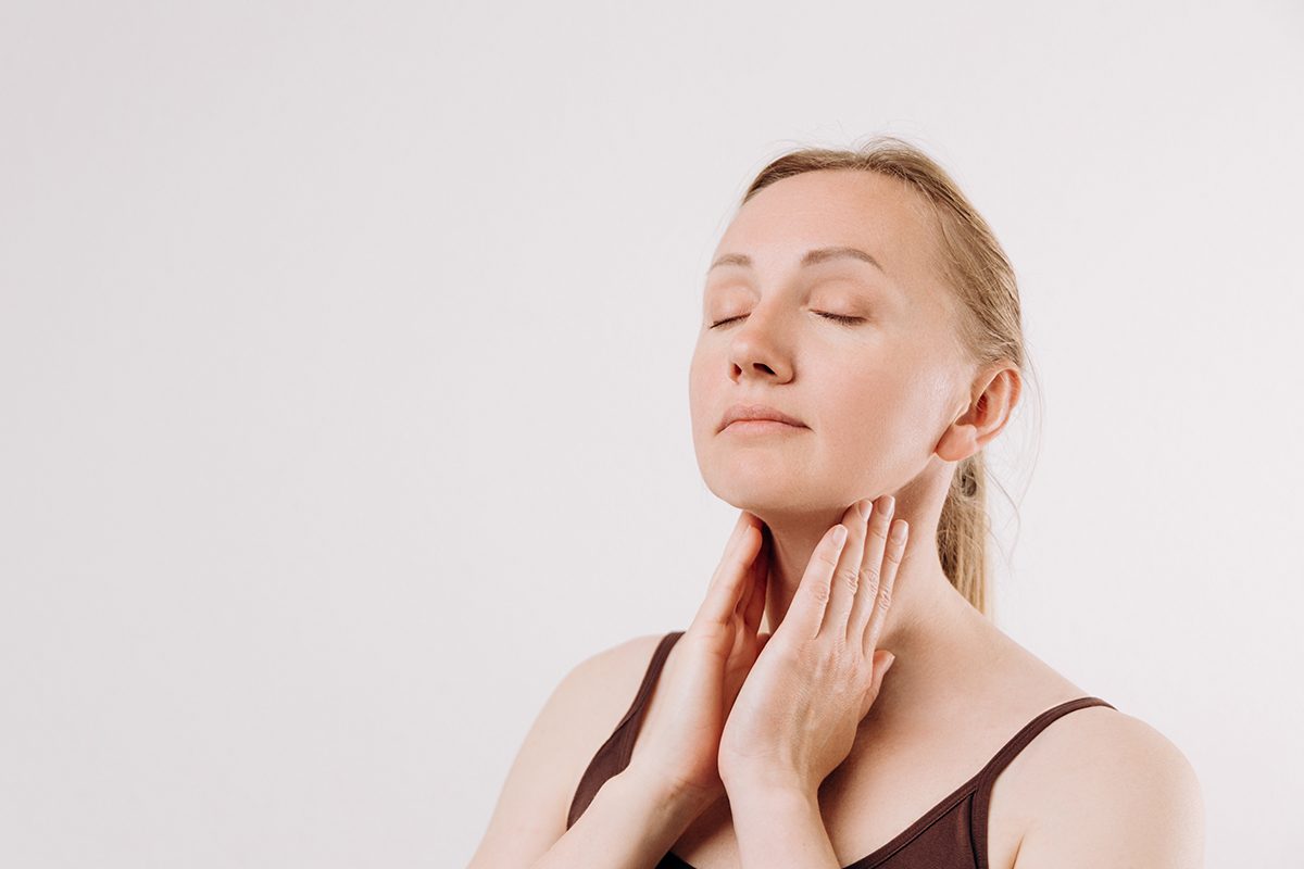 A woman stands with her eyes closed, gently touching her neck with both hands