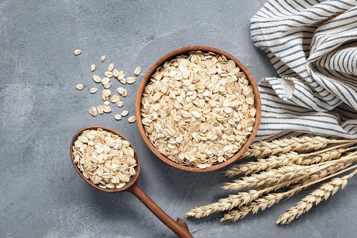 A wooden bowl and wooden spoon containing oats are set on a grey surface next to some stalks of wheat.