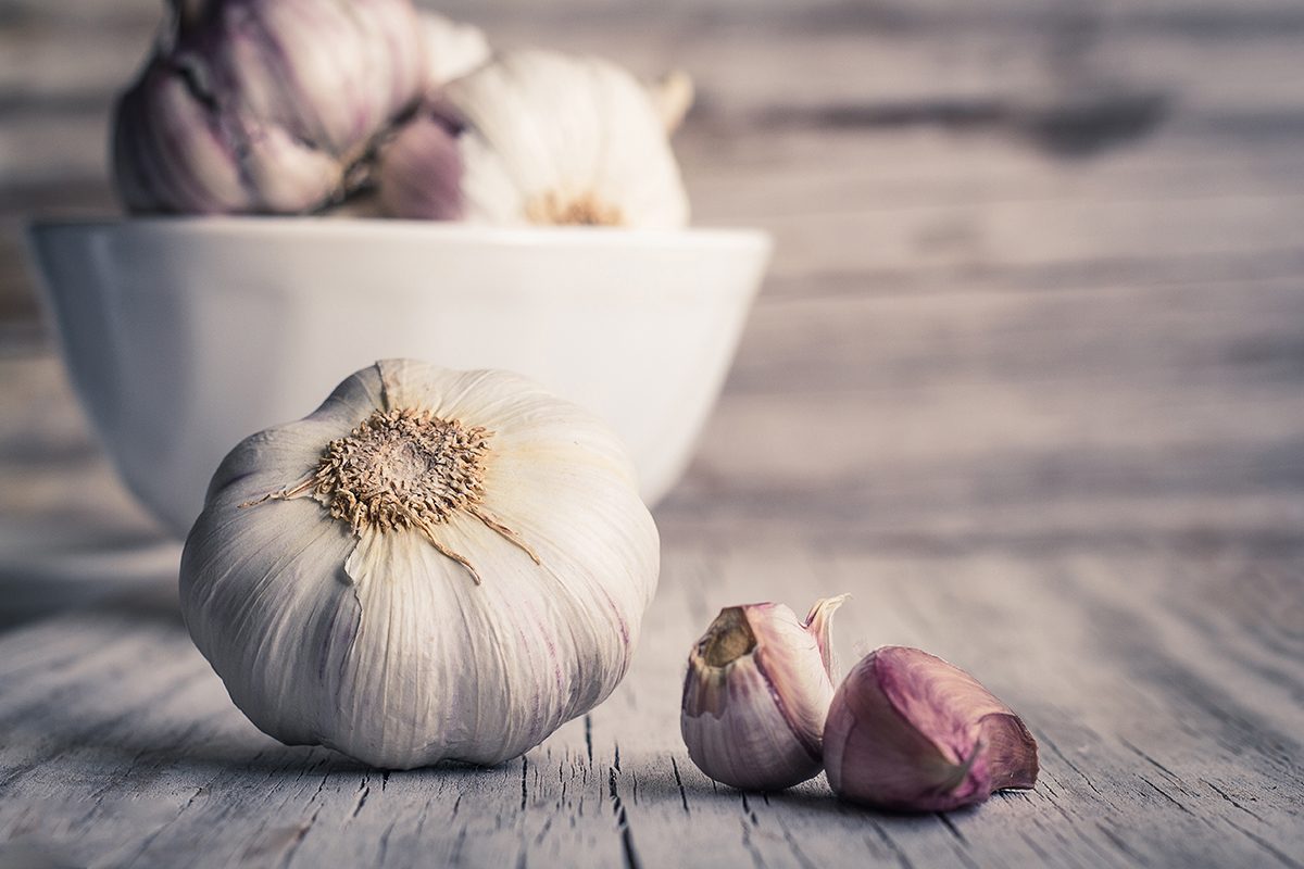 A garlic bulb and two cloves are on a wooden surface with a white bowl of garlic in the background.