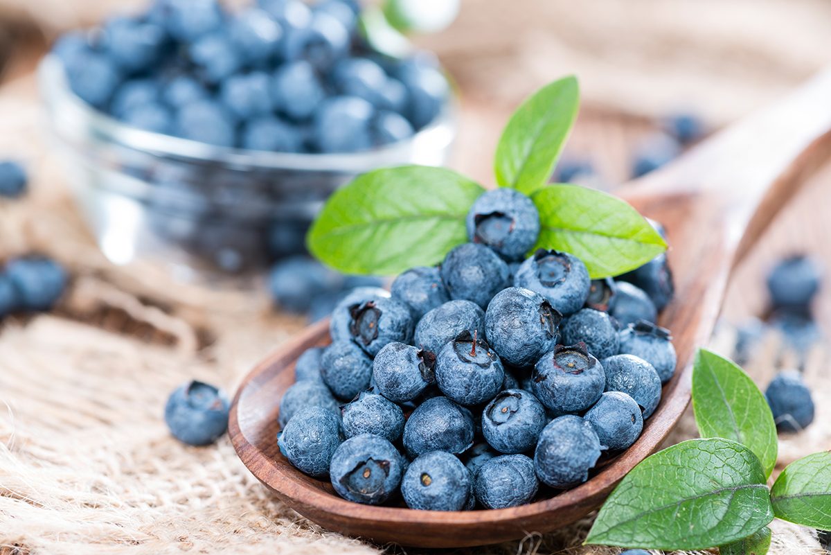 A wooden spoon with a heap of blueberries is positioned in front of a glass bowl, which also contains blueberries.