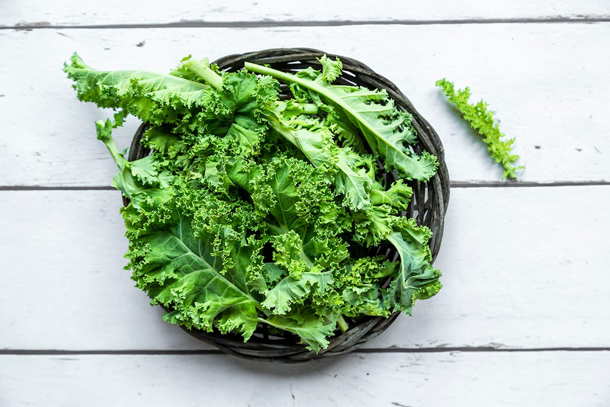 A small basket containing kale is on a white wooden surface.