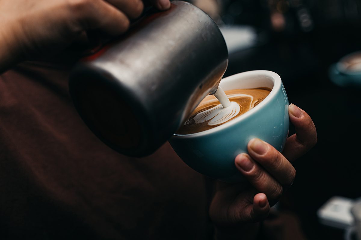 A person pours hot milk into a cup of coffee creating latte art.