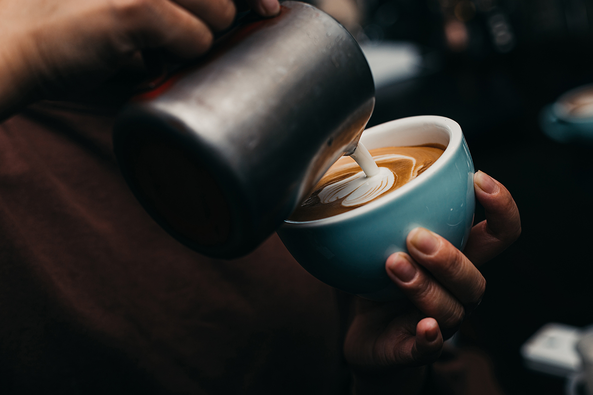 A person pours hot milk into a cup of coffee creating latte art.