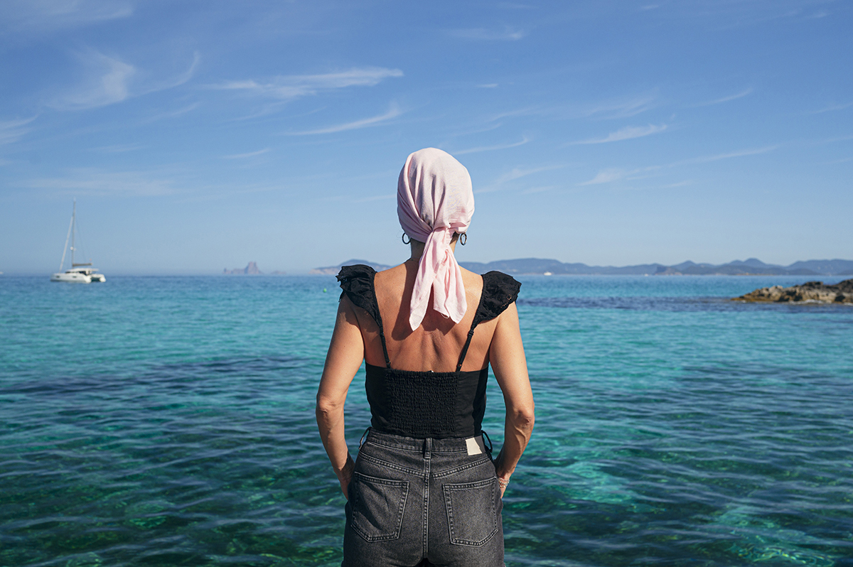 A woman wearing a pink headscarf looks out at a clear blue sea