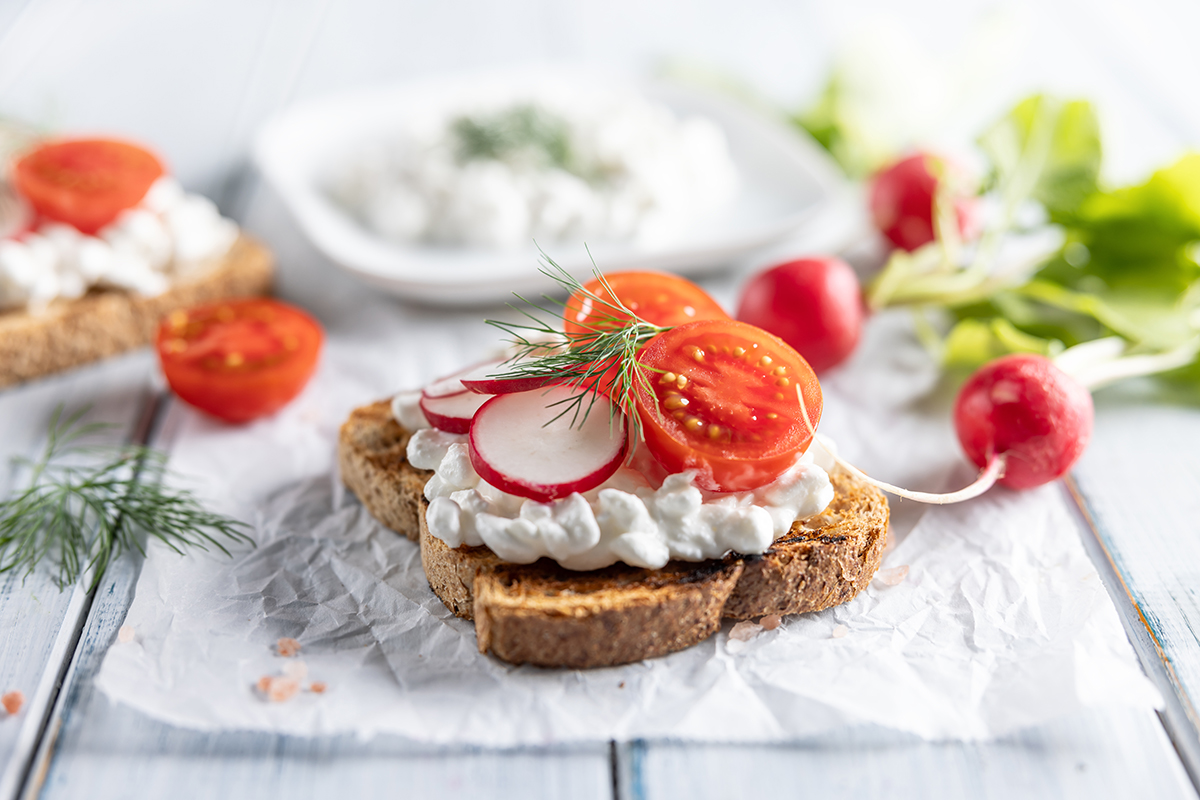 A toasted piece of brown bread with cottage cheese, a halved cherry tomato and sliced radish on top is set on a napkin on a wooden surface.
