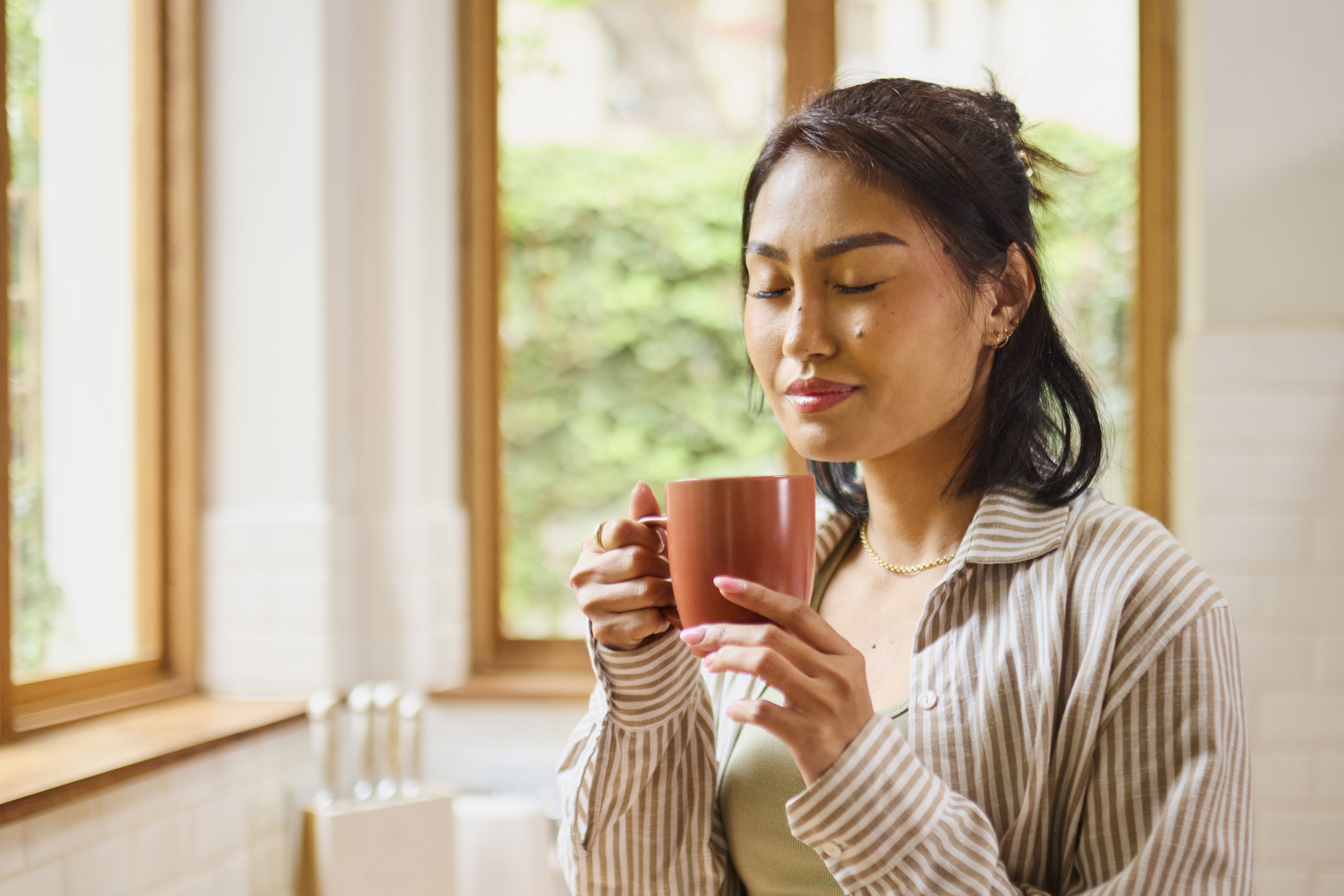 Person holding a ceramic mug near a window in a bright kitchen, with eyes closed and natural light illuminating the space.