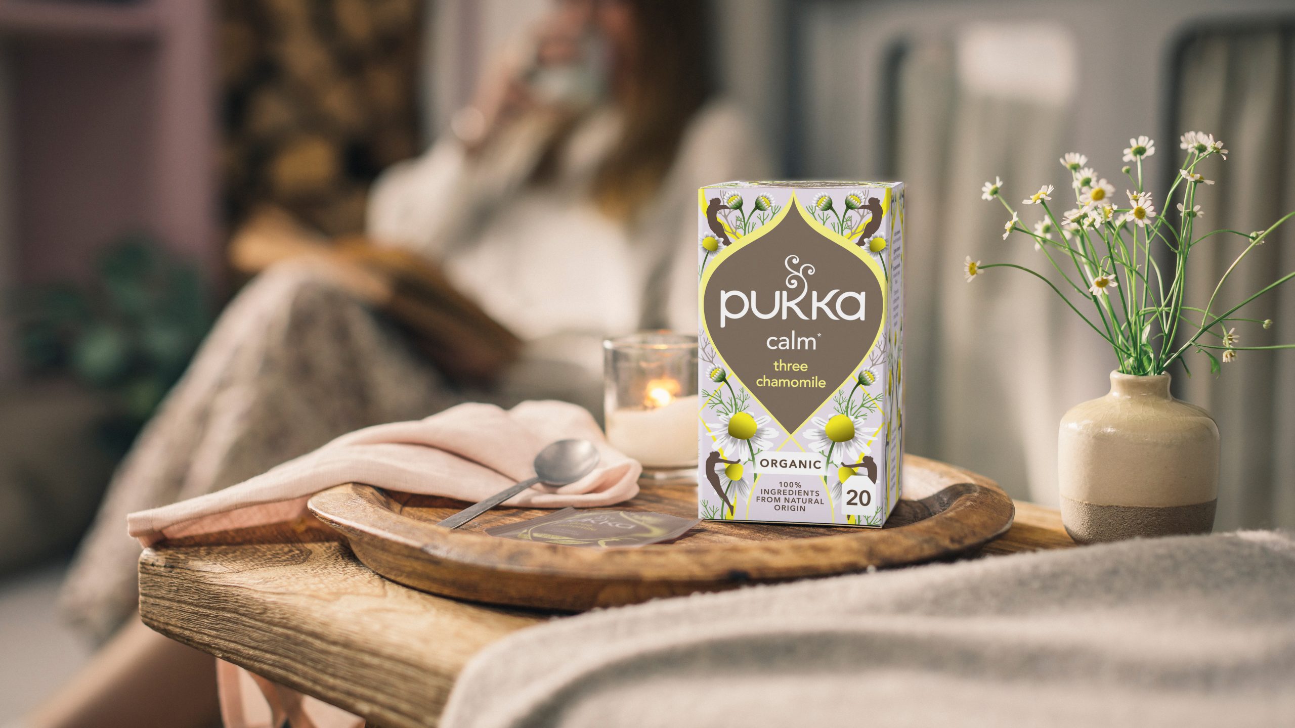 Box of Pukka Three Chamomile organic tea displayed on a wooden tray beside chamomile flowers and a candle in a home setting.