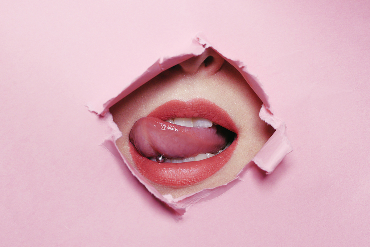 Pink paper with a hole in the middle reveals the mouth of a woman who is biting her tongue.