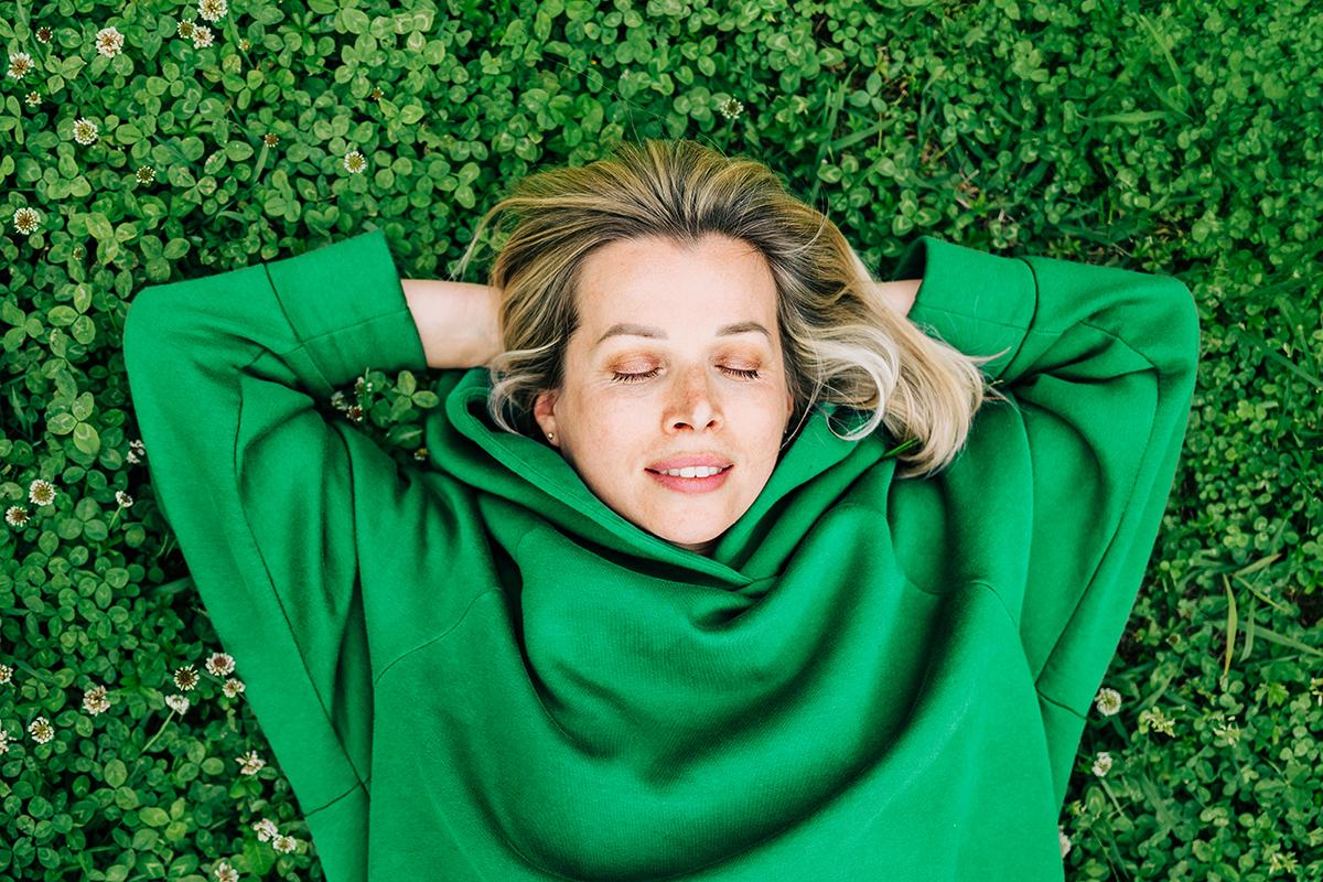 A woman wearing a green hoodie lies with her hands behind her head on a clover patch.