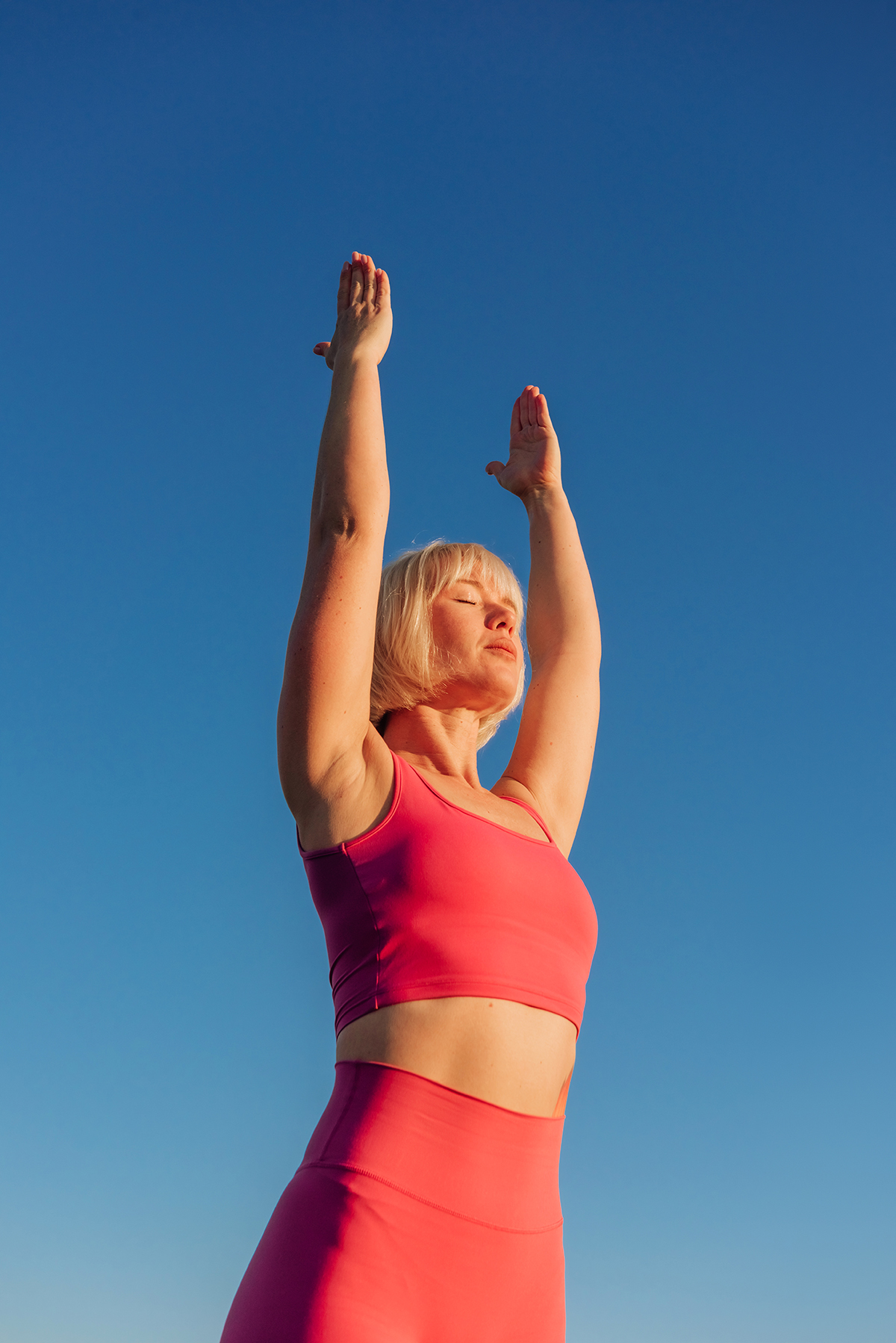 A blonde woman wearing a pink activewear set stretches her arms towards the sky with her eyes closed.
