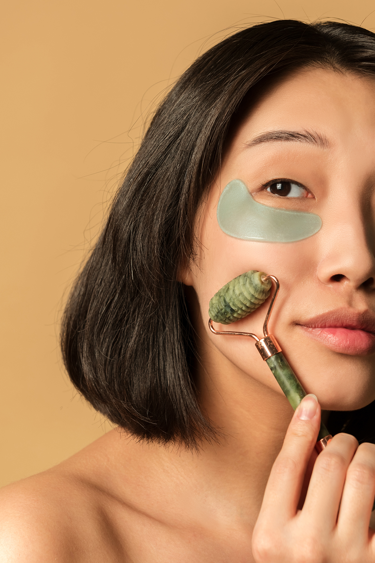 A close up portrait of a woman wearing an under eye patch rolling her cheek with a jade roller.