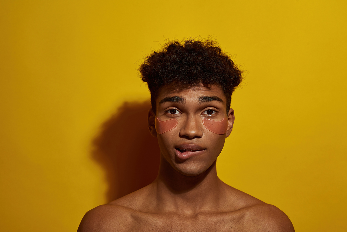 A man wearing under eye patches bites his lip in front of a yellow background.