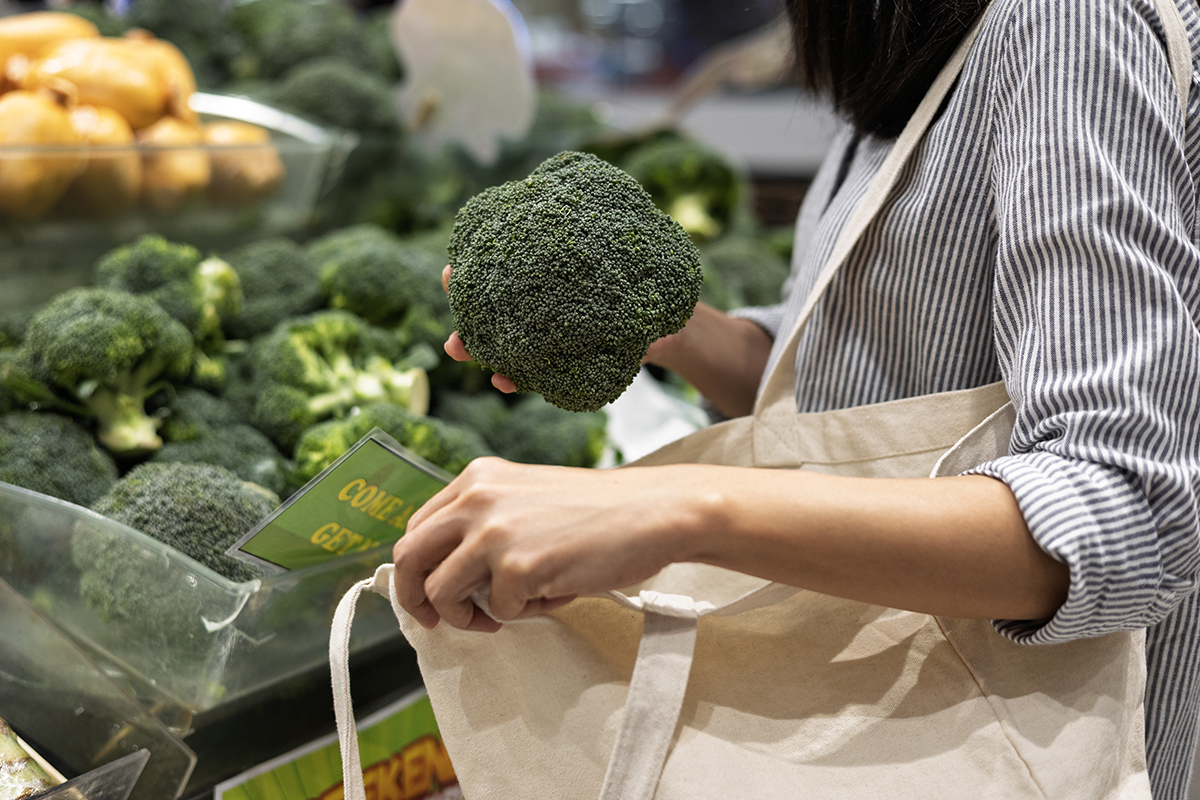 A woman is standing in a supermarket holding broccoli in one hand and opening a tote bag with the other.