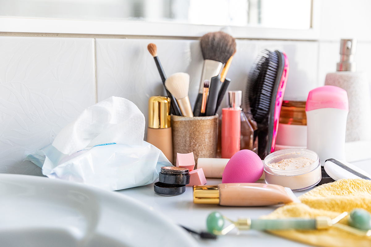 A range of makeup products, makeup brushes, wipes and a jade roller are strewn messily on a bathroom surface.