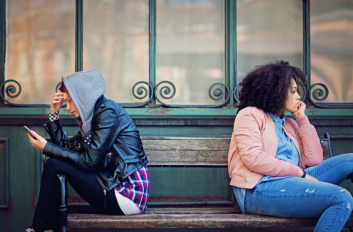 Two women are sat on a bench with their backs to each other and a gap in between them to signal separation. One of the women is looking at her phone and the other is staring ahead.