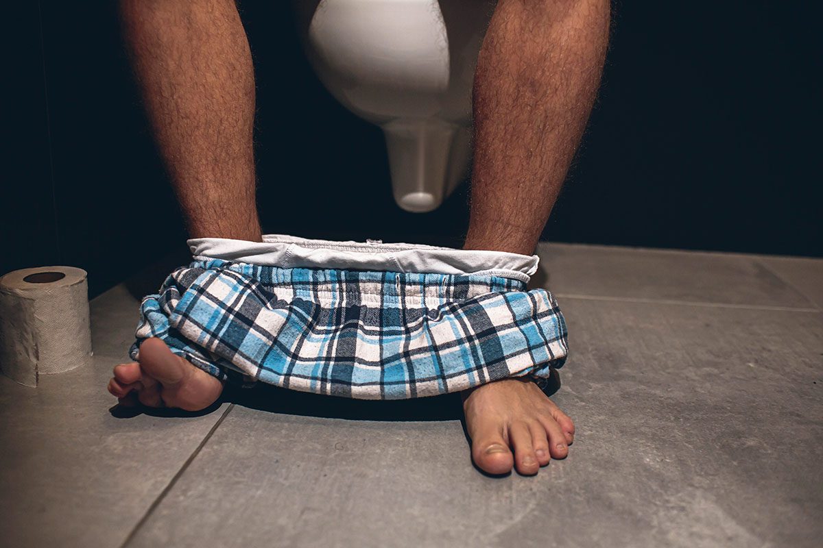A man sits on a toilet, with briefs around his ankles