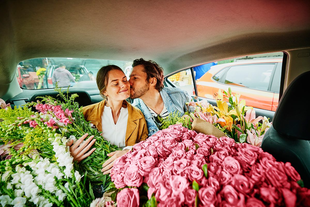 A couple are sitting in the back of a taxi, which is filled with flowers. The man is kissing the woman on the cheek.
