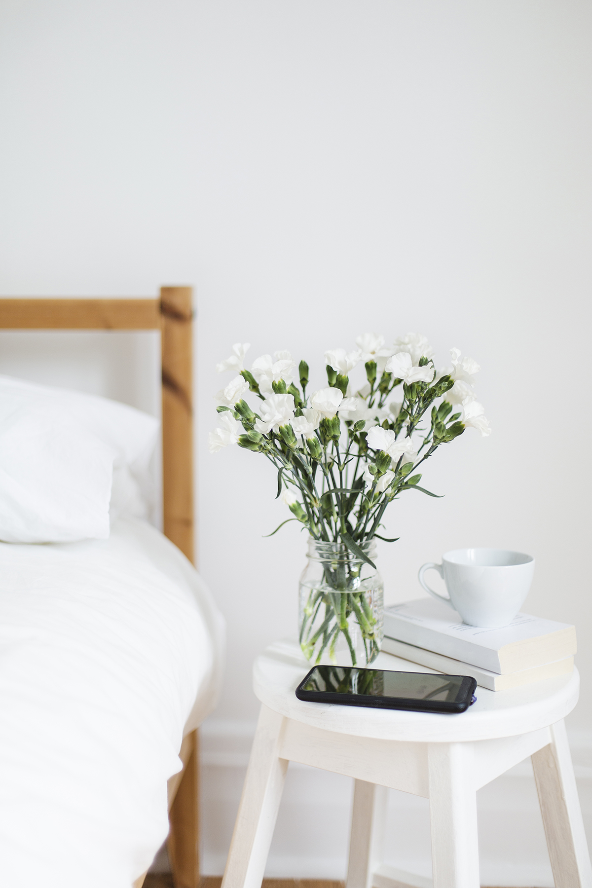A bouquet of white flowers are set on a white stool next to a bed.