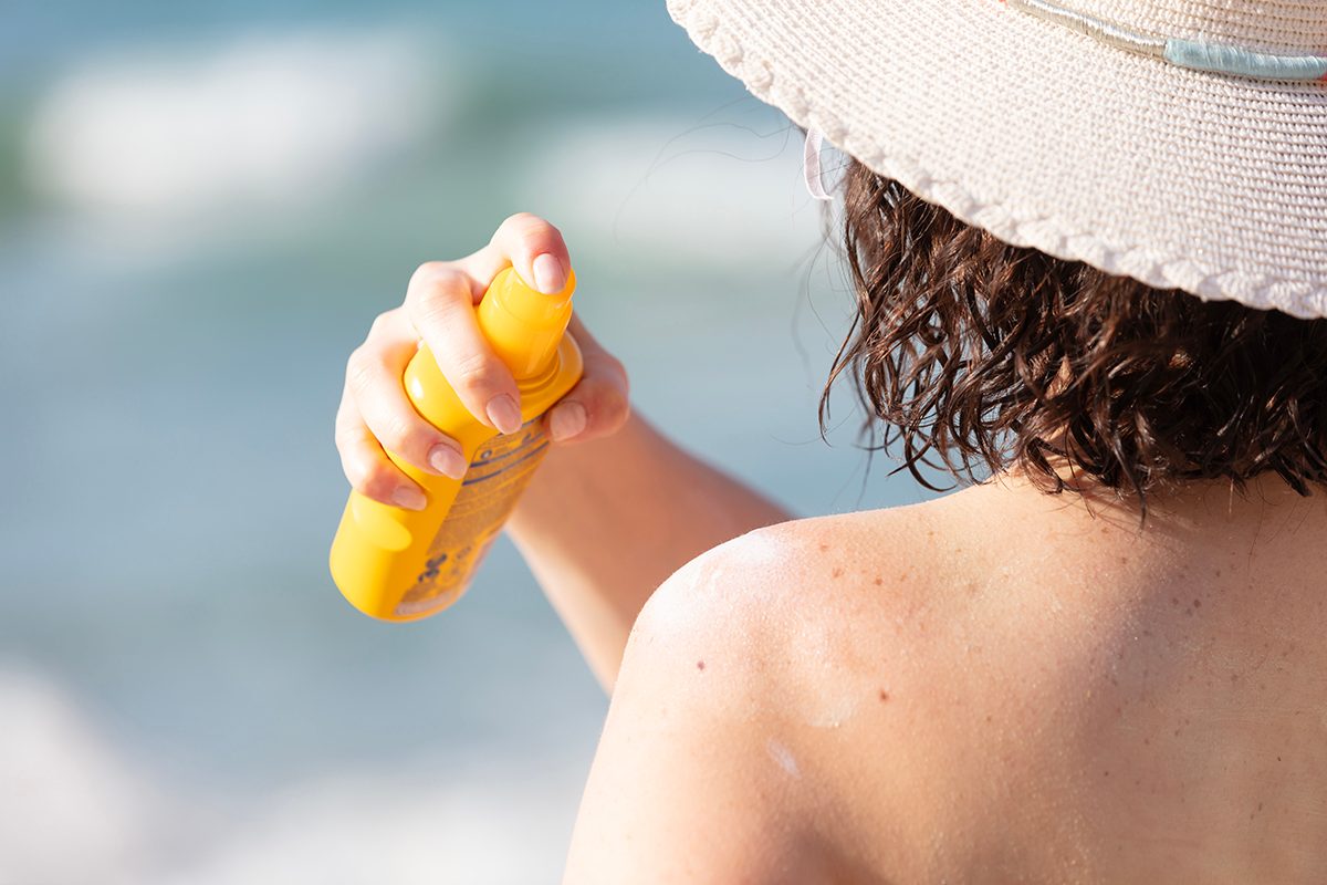 A woman wearing a sun hat sprays sunscreen onto her shoulder.