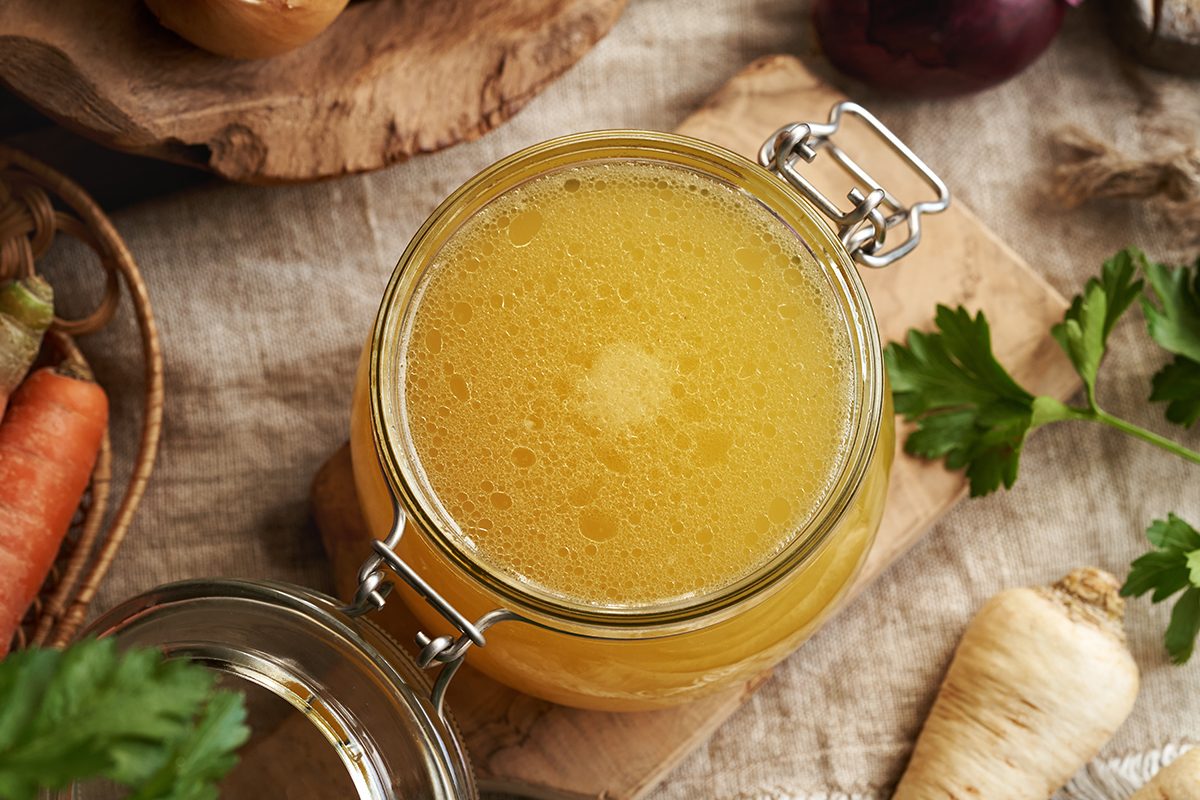 A glass jar filled with chicken bone broth is positioned on a wooden board, surrounded by fresh parsley, a carrot and a parsnip.