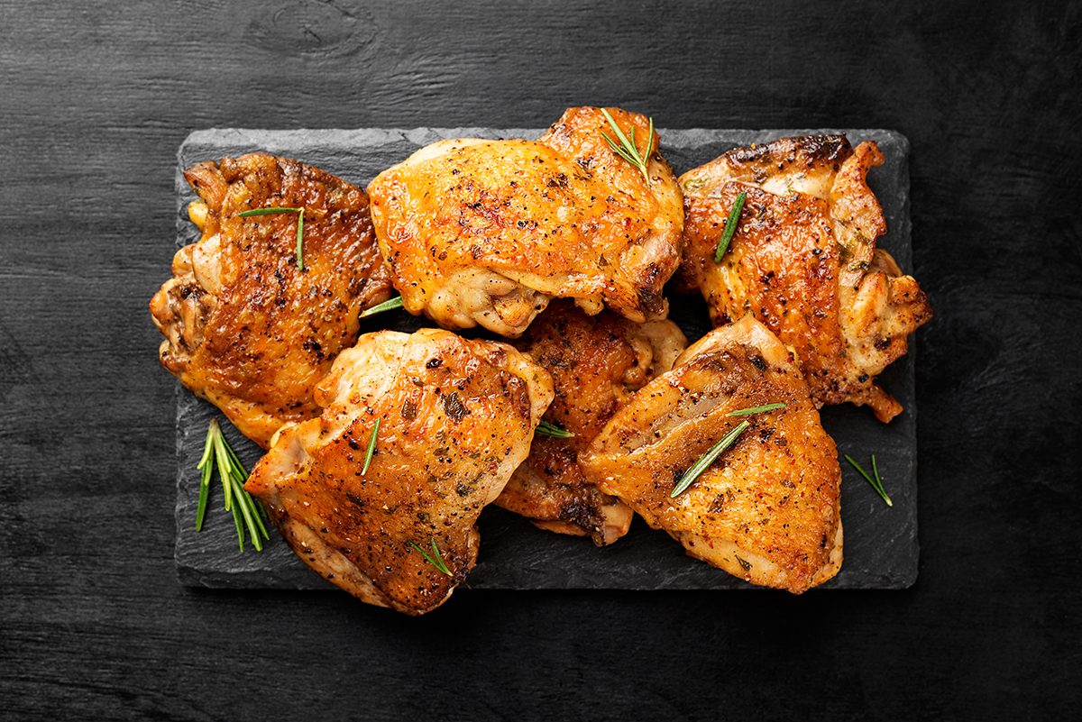 Cooked chicken thighs sprinkled with rosemary are positioned on a slate serving board, set against a black, wooden background. 