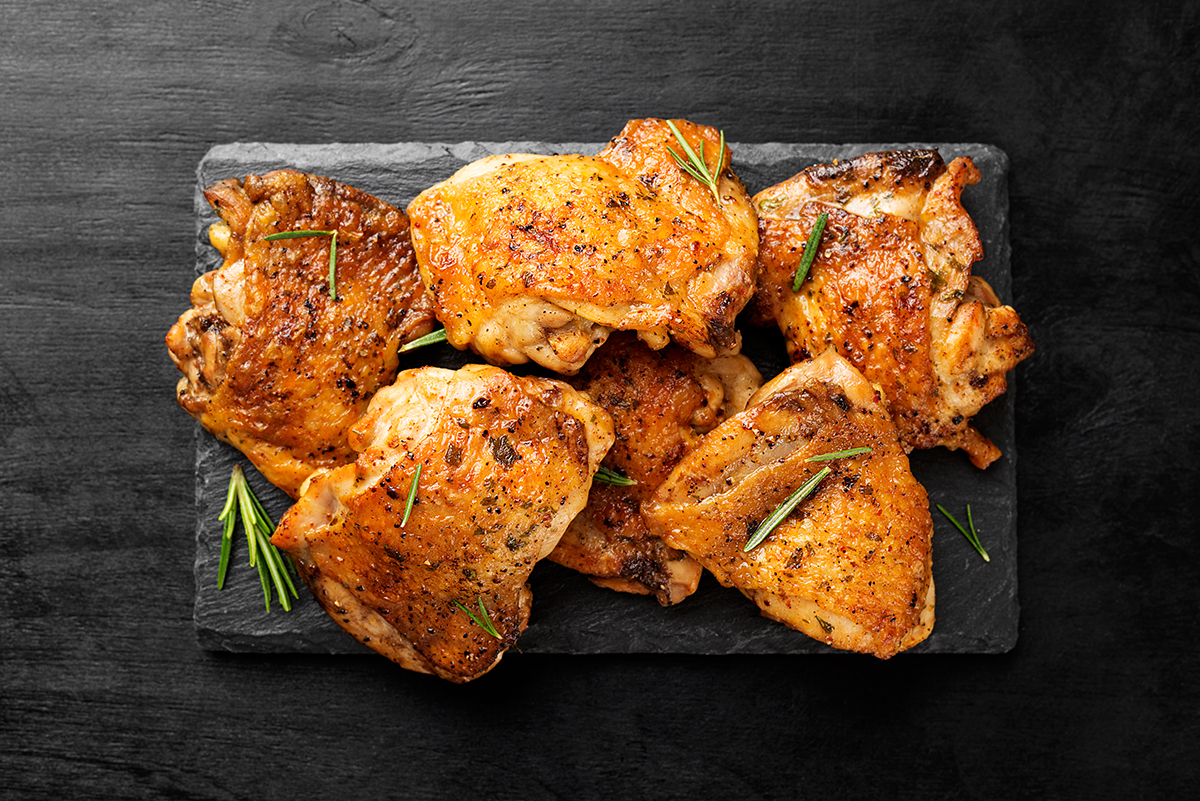 Cooked chicken thighs sprinkled with rosemary are positioned on a slate serving board, set against a black, wooden background. 