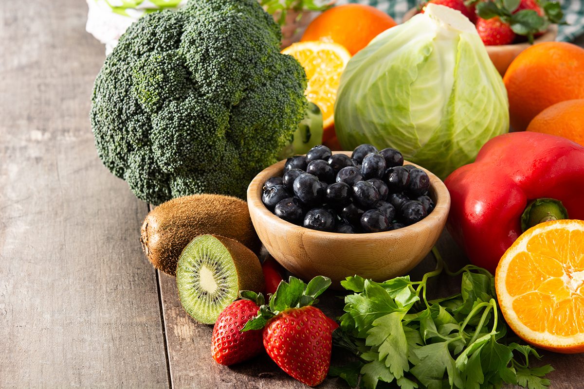 A selection of foods that are high in vitamin c sit on a wooden table, including broccoli, kiwis, strawberries, blueberries, a cabbage, a red pepper, oranges, and parsley. 