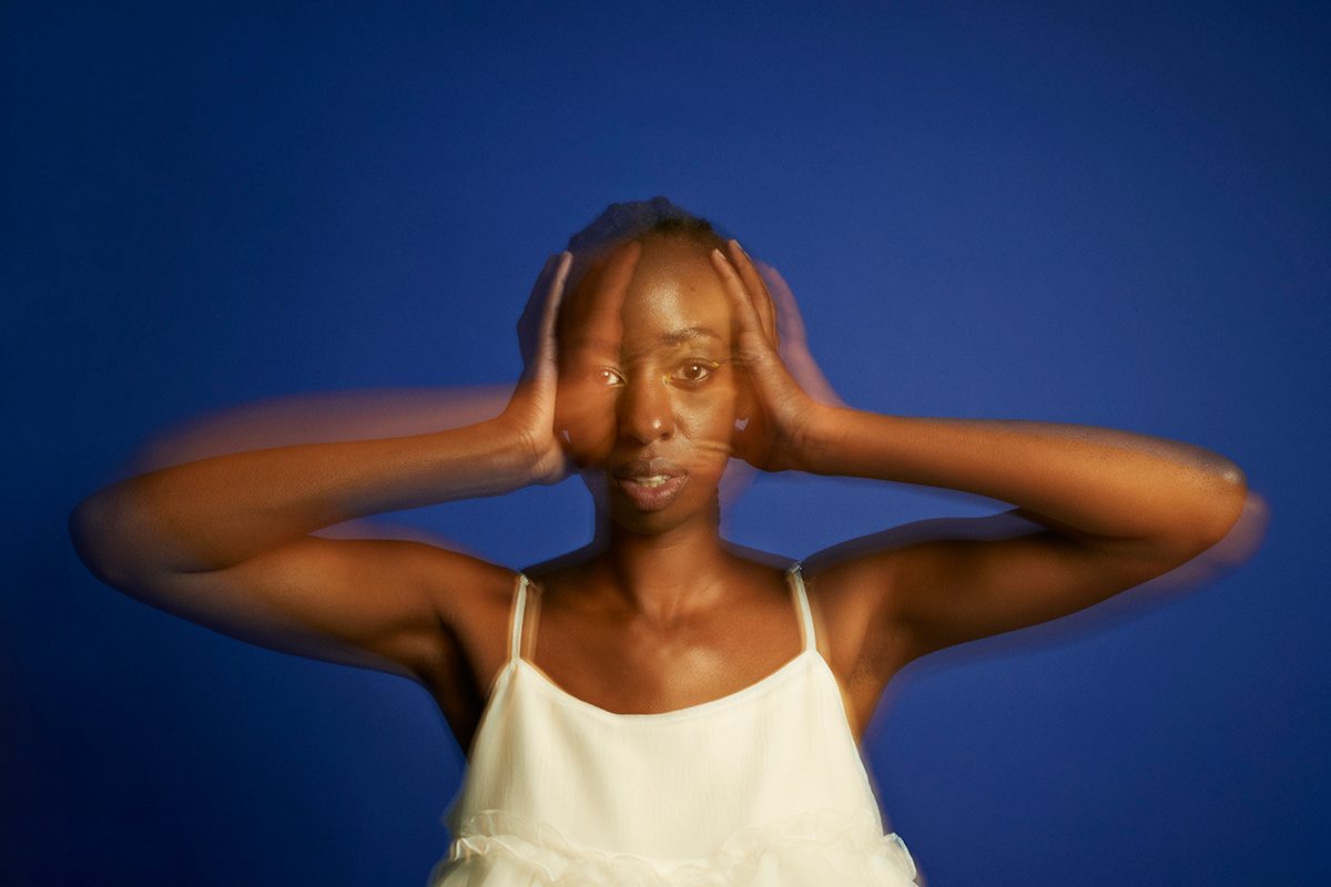 A blurred photo of a woman standing in front of a blue background, covering her ears with her hands