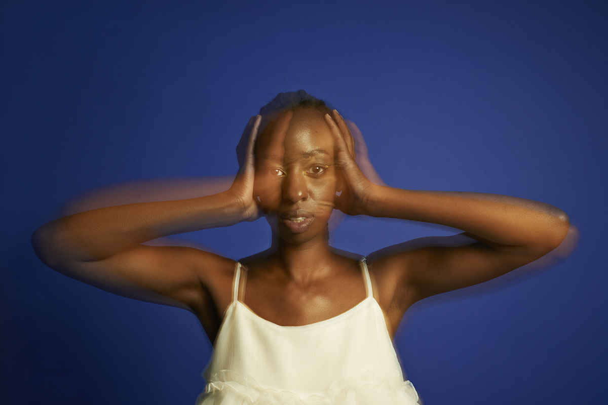 A blurred photo of a woman standing in front of a blue background, covering her ears with her hands