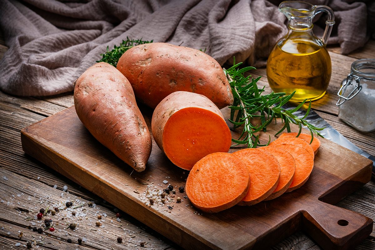 Whole and sliced raw sweet potatoes are positioned on a rustic wooden chopping board.