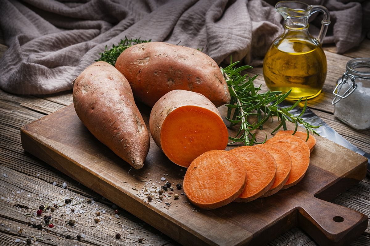 Whole and sliced raw sweet potatoes are positioned on a rustic wooden chopping board.