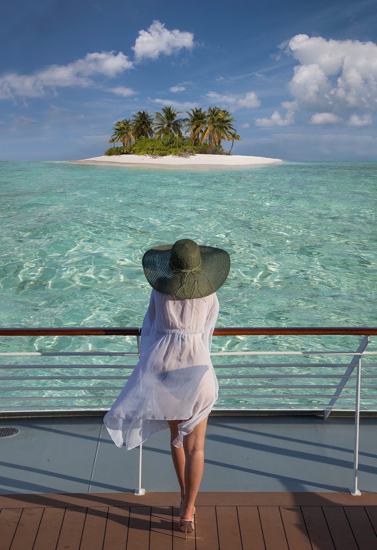 A woman stands on the deck of a cruise ship and looks out at a small island surrounded by clear water