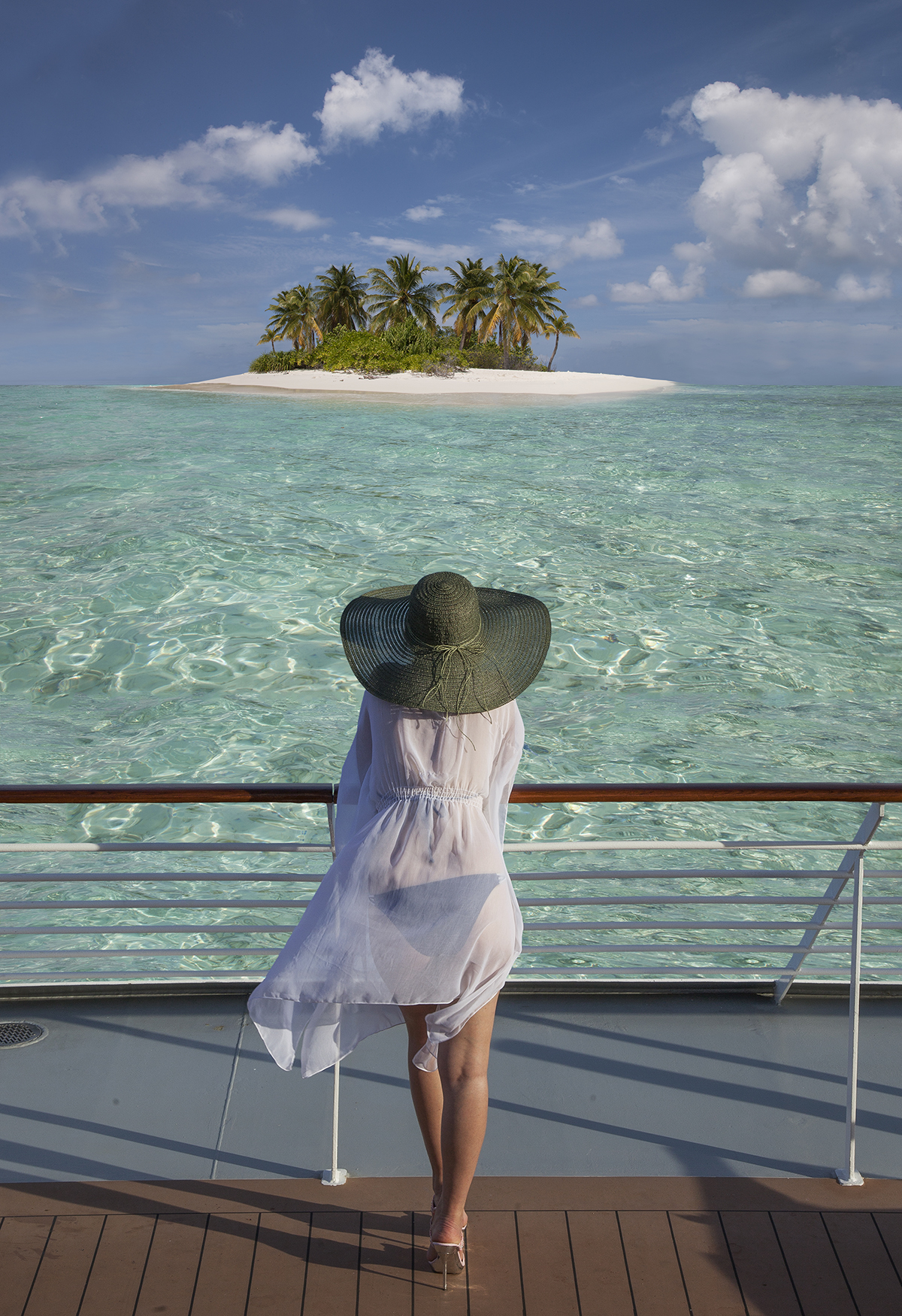A woman stands on the deck of a cruise ship and looks out at a small island surrounded by clear water